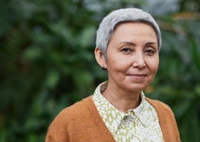 Portrait of an elegant senior Asian woman with a joyful expression, standing outdoors with greenery in the background.